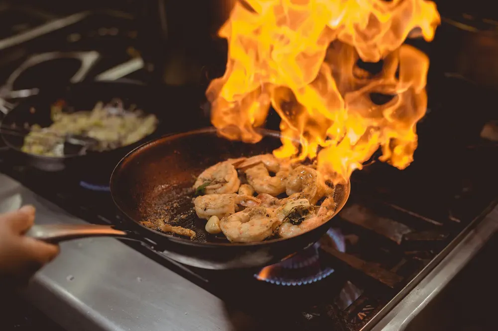 Close-up of a pan with shrimp sizzling while a wrapping flame emerges from the surface. A symbol of excessive heat on bone cells.